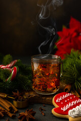 Glass mug with brewed black tea with steam among spruce branches and Christmas Poinsetia flower, gingerbread in the form of a red mitten and sticks candy, cozy festive winter evening