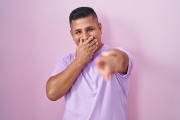 Young hispanic man standing over pink background laughing at you, pointing finger to the camera...