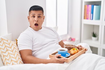 Young hispanic man eating breakfast in the bed scared and amazed with open mouth for surprise,...
