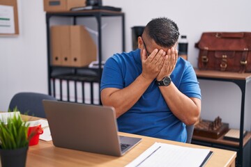 Young hispanic man working at the office with laptop with sad expression covering face with hands...