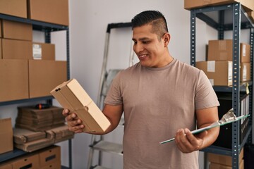 Young latin man ecommerce business worker reading document holding package at office
