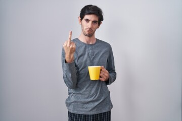 Young hispanic man wearing pajama drinking a cup of coffee showing middle finger, impolite and rude...