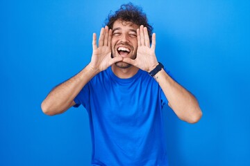 Hispanic young man standing over blue background smiling cheerful playing peek a boo with hands showing face. surprised and exited