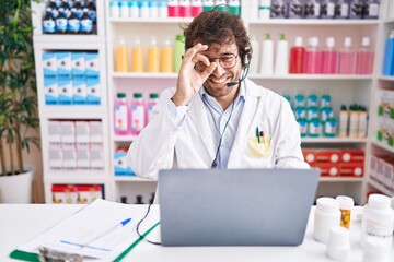 Hispanic young man working at pharmacy drugstore working with laptop smiling happy doing ok sign with hand on eye looking through fingers