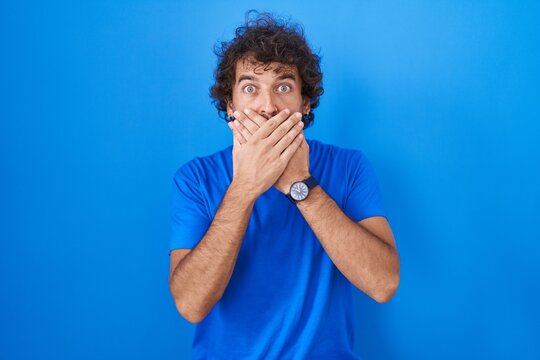 Hispanic young man standing over blue background shocked covering mouth with hands for mistake. secret concept.