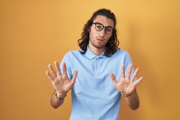 Young hispanic man standing over yellow background moving away hands palms showing refusal and...
