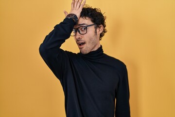 Hispanic man standing over yellow background surprised with hand on head for mistake, remember...