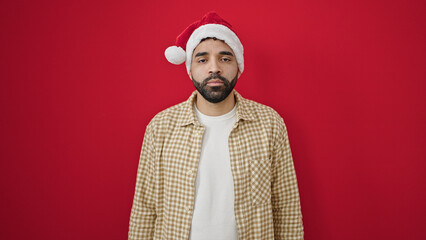 Young hispanic man standing with serious expression wearing christmas hat over isolated red background