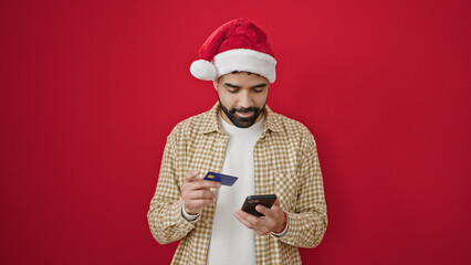 Young hispanic man shopping with smartphone and credit card wearing christmas hat over isolated red background