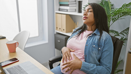 Young successful hispanic businesswoman, pregnant and calmly massaging her belly while taking a...