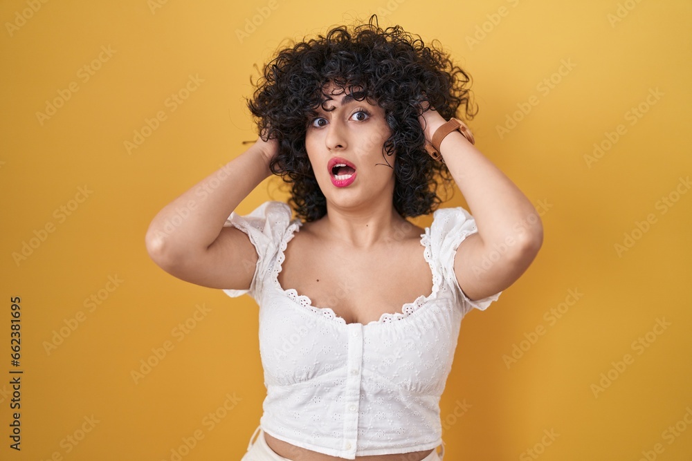 Poster young brunette woman with curly hair standing over yellow background crazy and scared with hands on 