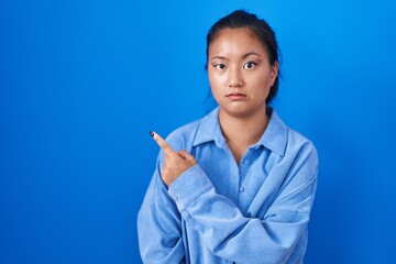 Asian young woman standing over blue background pointing aside worried and nervous with forefinger,...