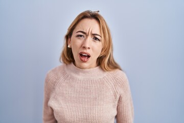 Hispanic woman standing over blue background in shock face, looking skeptical and sarcastic,...