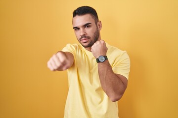 Young hispanic man standing over yellow background punching fist to fight, aggressive and angry...