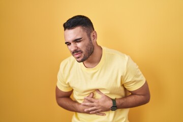 Young hispanic man standing over yellow background with hand on stomach because indigestion, painful illness feeling unwell. ache concept.