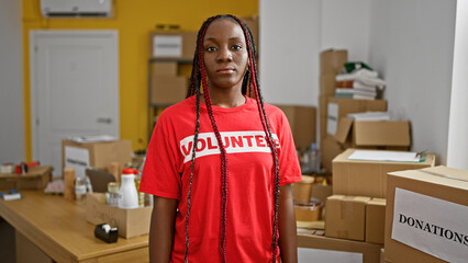 Beautiful african american woman, a serious-faced volunteer, stands indoors in charity center's...
