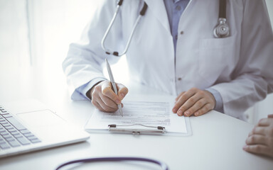 Doctor and patient sitting at the table in clinic. The focus is on female physician's hands filling up the medication history record form or checklist, close up. Medicine concept