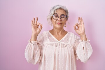 Middle age woman with grey hair standing over pink background relax and smiling with eyes closed doing meditation gesture with fingers. yoga concept.