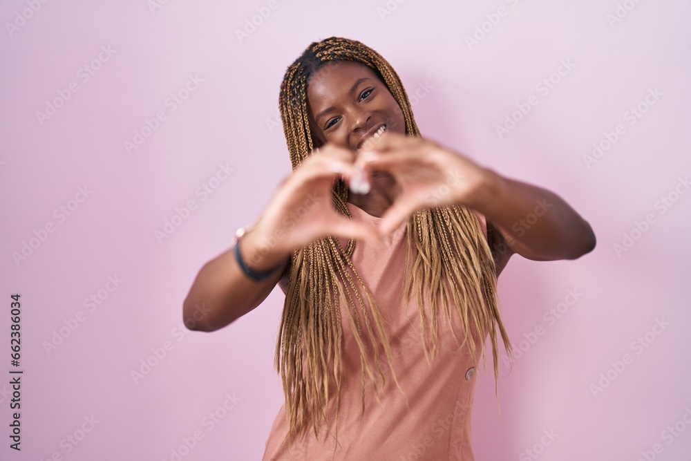 Wall mural African american woman with braided hair standing over pink background smiling in love doing heart symbol shape with hands. romantic concept.