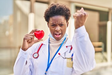 African american woman wearing doctor uniform holding heart annoyed and frustrated shouting with anger, yelling crazy with anger and hand raised