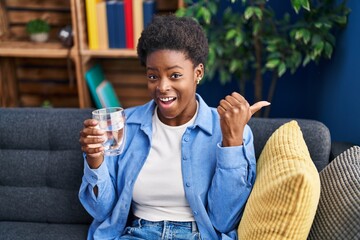 African american woman drinking glass of water pointing thumb up to the side smiling happy with...