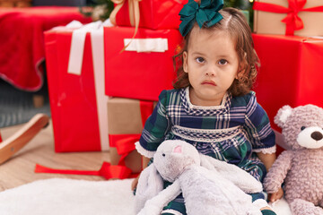 Adorable blonde toddler sitting on floor by christmas gifts with relaxed expression at home