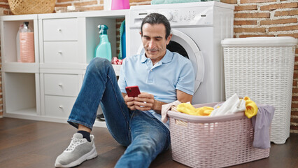 Middle age man using smartphone waiting for washing machine at laundry room