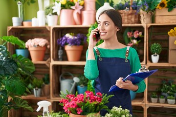 Young beautiful hispanic woman florist talking on smartphone reading document at flower shop