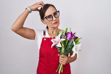 Middle age brunette woman wearing apron working at florist shop holding bouquet confuse and...