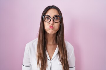Young brunette woman wearing glasses standing over pink background puffing cheeks with funny face. mouth inflated with air, crazy expression.