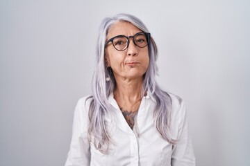 Middle age woman with tattoos wearing glasses standing over white background puffing cheeks with funny face. mouth inflated with air, crazy expression.