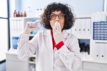 Hispanic doctor woman with curly hair holding vaccine covering mouth with hand, shocked and afraid...