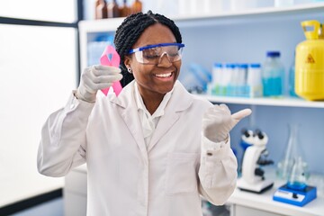 Naklejka premium African american woman working at scientist laboratory holding pink ribbon pointing thumb up to the side smiling happy with open mouth
