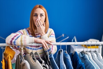 Young woman searching clothes on clothing rack covering mouth with hand, shocked and afraid for...