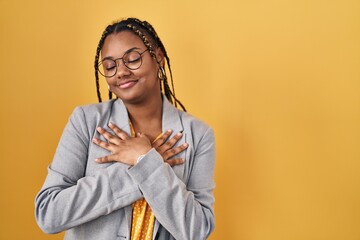 African american woman with braids standing over yellow background smiling with hands on chest with closed eyes and grateful gesture on face. health concept.