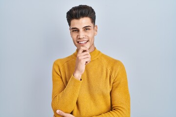 Young hispanic man standing over blue background looking confident at the camera smiling with crossed arms and hand raised on chin. thinking positive.