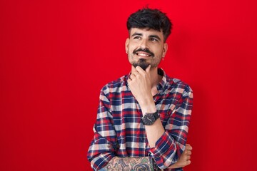 Young hispanic man with beard standing over red background with hand on chin thinking about question, pensive expression. smiling and thoughtful face. doubt concept.