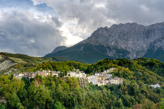Quaint hillside village of Castelli in Abruzzo, nestled against majestic mountains. Sunlight bathes the historic homes surrounded by lush greenery under a dramatic sky.