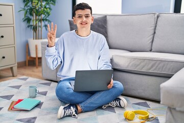 Non binary person studying using computer laptop sitting on the floor showing and pointing up with fingers number three while smiling confident and happy.