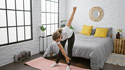 Serious young blonde woman finding balance, strengthening her concentration with morning yoga exercise on her bed, warming up in cozy indoor bedroom space, enjoying calmness and relaxation. - obrazy, fototapety, plakaty