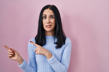 Hispanic woman standing over pink background pointing aside worried and nervous with both hands,...