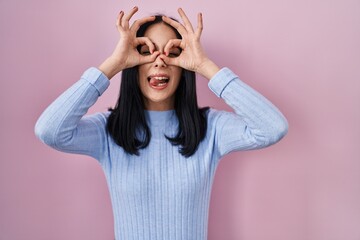 Hispanic woman standing over pink background doing ok gesture like binoculars sticking tongue out,...