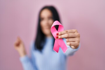 Hispanic woman holding pink cancer ribbon smiling happy pointing with hand and finger to the side