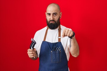 Young hispanic man with beard and tattoos wearing barber apron holding razor pointing down looking...