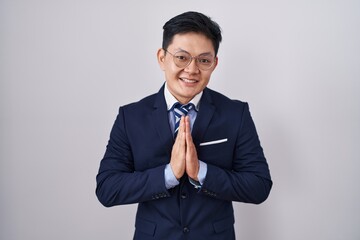 Young asian man wearing business suit and tie praying with hands together asking for forgiveness smiling confident.
