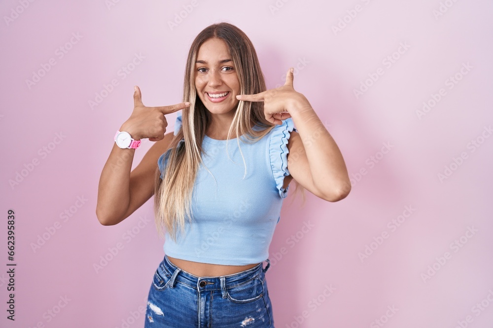 Poster Young blonde woman standing over pink background smiling cheerful showing and pointing with fingers teeth and mouth. dental health concept.