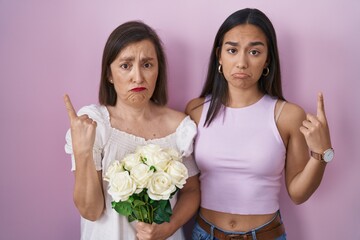 Hispanic mother and daughter holding bouquet of white flowers pointing up looking sad and upset, indicating direction with fingers, unhappy and depressed.