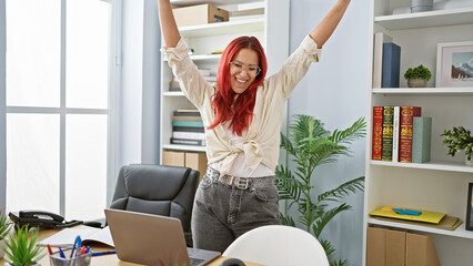 Joyful young redhead woman worker celebrating business win at office, confidently working on laptop at desk, positive professional energy indoors.