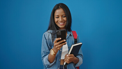 African american woman student using smartphone holding book over isolated blue background