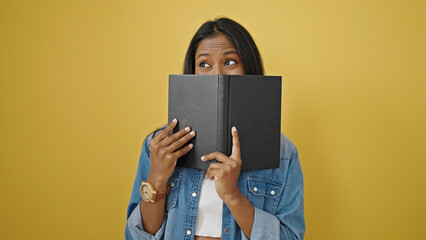 African american woman covering mouth with book over isolated yellow background
